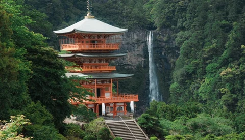 Red-and-gold pagoda with multiple tiers nestled against a tall waterfall in a forested canyon.