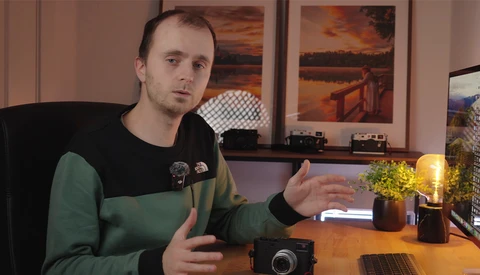 Man in green and black shirt gesturing while speaking at desk with camera and laptop.