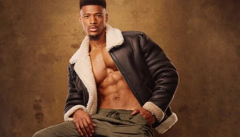 Shirtless man in black leather jacket with sherpa collar posing against textured brown backdrop.