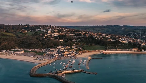Coastal town with curved harbor pier at golden hour, surrounded by hillside homes and sandy beach.