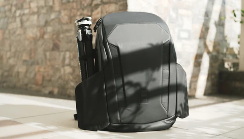 Black hard-shell camera backpack photographed outdoors with natural sunlight creating strong shadows on pavement.