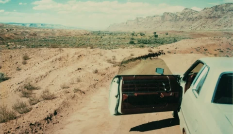 White vehicle with open door parked on desert road overlooking vast arid landscape with scattered vegetation.