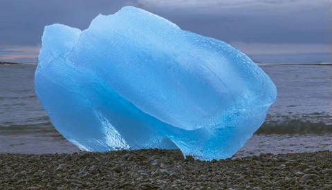 Luminous blue iceberg resting on a rocky beach against a twilight sky.