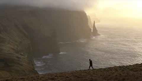 Solitary hiker walking on a cliff edge overlooking dramatic coastal rock formations shrouded in mist at sunrise.