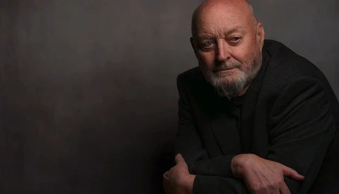 Bald man with beard in dark clothing against neutral gray background.