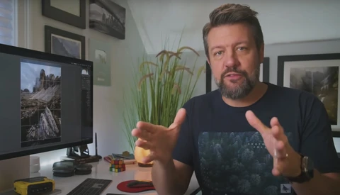 Man gesturing while discussing photography at a desk with a monitor displaying landscape imagery.