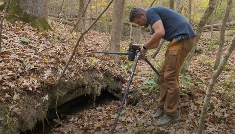 Man using a metal detector in a wooded area covered with fallen leaves.