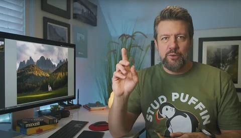 Man in green shirt gesturing while speaking at a desk with a monitor displaying mountain landscape photography.