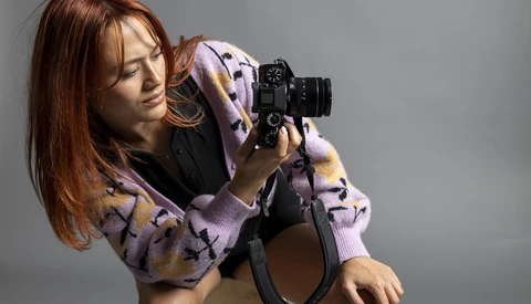 Woman holding a film camera to her eye, wearing a lavender patterned cardigan against a neutral gray background.
