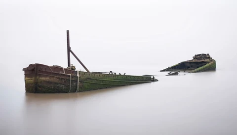 Two rusted, abandoned shipwrecks partially submerged in still water with algae-covered hulls.