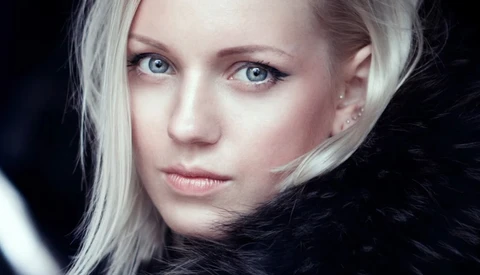Woman with platinum blonde hair and blue eyes gazing toward camera against dark background.