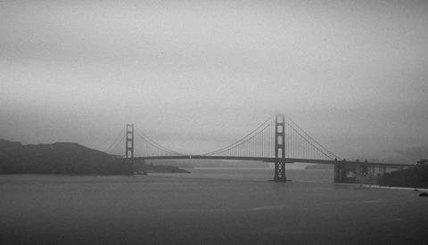 Suspension bridge spanning a body of water under overcast skies in black and white.