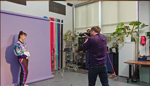 Cinematographer filming a portrait subject against a purple backdrop in a studio with professional lighting equipment.