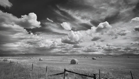 Expansive prairie landscape with wooden ranch fence in foreground and dramatic cloudy sky overhead.