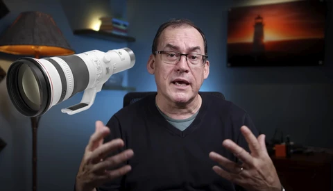 Man wearing glasses speaking to camera with professional telephoto lens visible in foreground.
