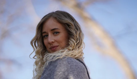 Portrait of a woman with wavy blonde hair wearing a gray sweater, looking over her shoulder against a blurred blue sky background.