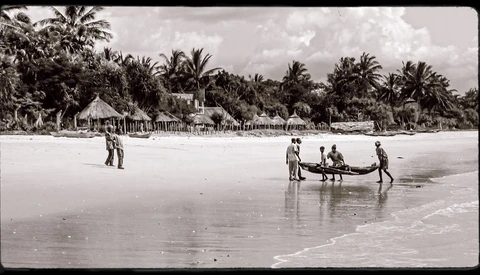 Multiple figures launching a wooden boat into shallow ocean waters with palm-lined beach village visible in background.