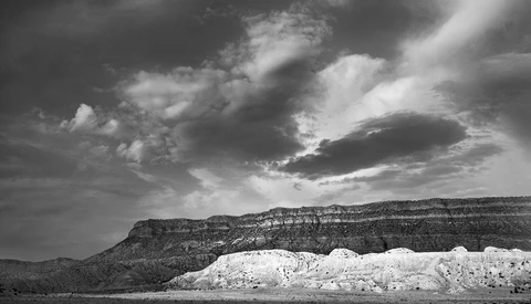 Dramatic black and white landscape with layered mesa formations and white cliff face under stormy sky.