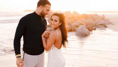 Couple embracing on a beach at sunset with rocky formations in the background.