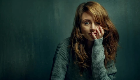 Woman with long brown hair resting her face against her hand, wearing a gray sweater against a teal background.