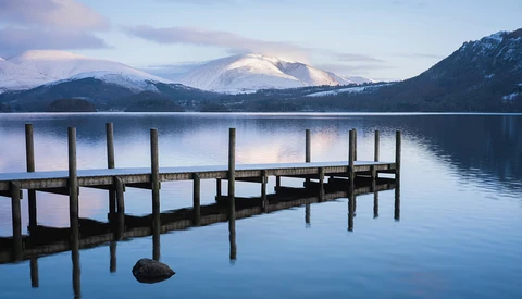 Wooden dock extending into still lake with snow-capped mountains reflected in water at dawn.