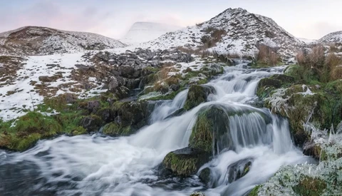 Cascading waterfall flowing over moss-covered rocks with snow-dusted mountains in the background.