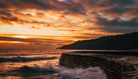 Dramatic coastal sunset with layered rock formations in foreground and mountainous headland silhouetted against golden sky.