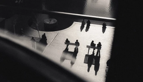 Overhead view of silhouetted figures casting long shadows on a bright floor beneath architectural elements.
