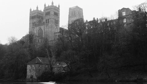 Gothic cathedral with twin spires rises above bare trees and a riverside building in monochromatic tones.