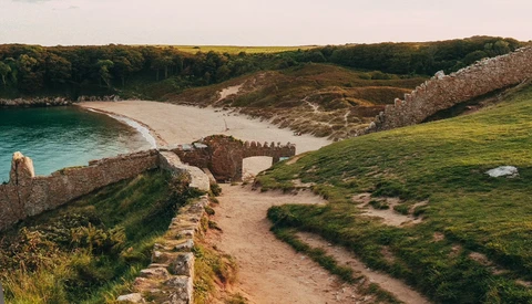 Coastal landscape with turquoise water, sandy beach, stone ruins, and green hillside covered in grass.