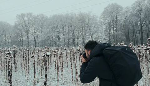 Photographer in black jacket shooting a frost-covered cornfield with bare trees in misty background.