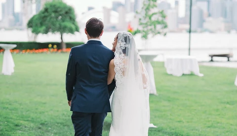 Bride and groom walking together on a grassy venue with a city skyline in the background.