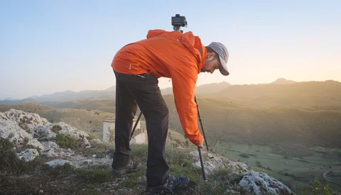 Hiker in orange jacket bending forward on rocky hilltop, adjusting camera mounted on head strap.