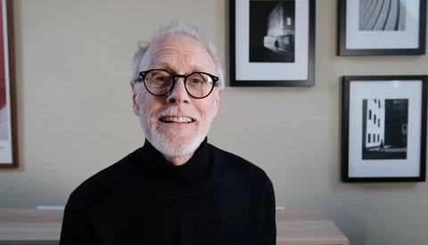 Older man with white hair and glasses wearing black turtleneck, seated indoors with framed black and white architectural photographs on wall behind.