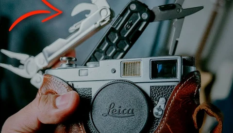 Photographer's hands holding a Leica rangefinder camera with brown leather strap, surrounded by vintage camera accessories and film photography gear.