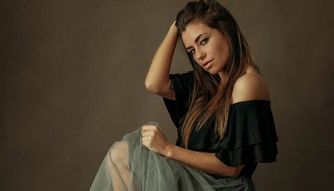 Woman in off-shoulder black top and gray skirt poses against neutral backdrop with hand to head.