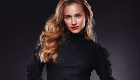 Woman with long wavy golden-blonde hair wearing a black turtleneck against a dark background.