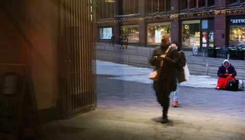 Woman in dark coat walking across an urban plaza at dusk, storefront windows visible in background.