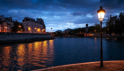 Illuminated riverside cityscape at twilight with ornate street lamp in foreground.