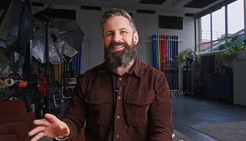 Bearded man in brown shirt gesturing while speaking in a photography studio with equipment and large windows.
