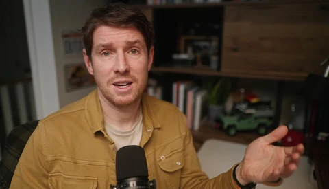 Man in mustard shirt speaking into microphone during interview or podcast recording.