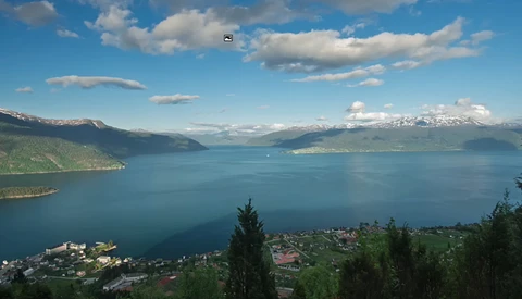 Alpine lake surrounded by forested mountains under partly cloudy sky with small settlements on shoreline.