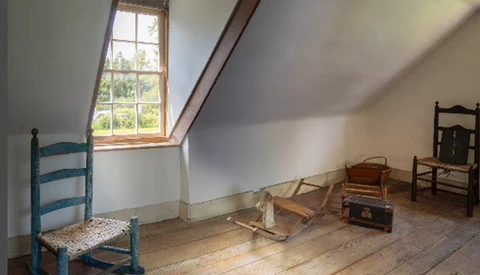 Sparse attic room with sloped ceiling, tall window, wooden floorboards, and mismatched vintage chairs and furnishings.