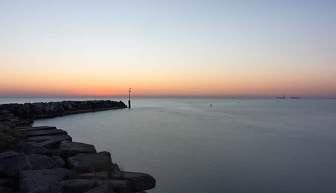 Rocky jetty extending into calm water at twilight with a solitary navigation marker visible in the distance.
