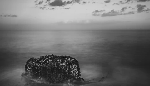 Weathered lobster trap partially submerged in shallow ocean water under overcast sky.