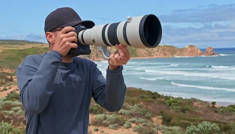 Photographer with telephoto lens positioned on coastal bluff overlooking rocky headland and ocean.
