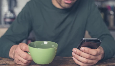 Person holding a green mug and smartphone while seated at a table, wearing a dark long-sleeved shirt.