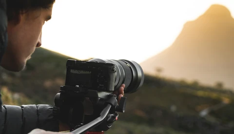 Photographer operating a mirrorless camera on a tripod during golden hour with mountains in the background.