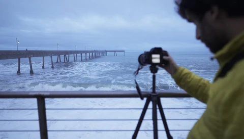 Photographer in yellow jacket capturing pier extending into ocean at twilight using tripod-mounted camera.