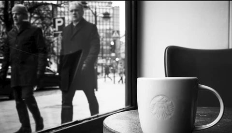 White ceramic mug with emblem on table beside window, blurred figures visible on street outside.